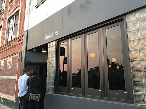 The entrance to a dark storefront restaurant at dusk, the name spelled out in gold script above glass block windows and paneled doors.