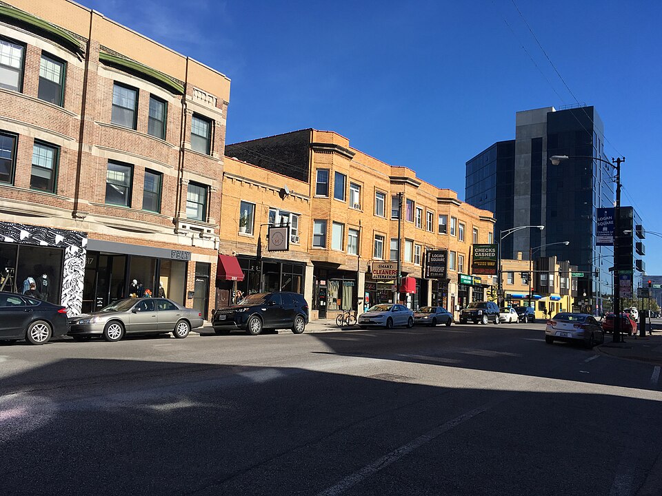 A Milwaukee Avenue corner with a Logan Square street banner on the lamppost, older brick buildings with apartments over the shops next to a newer glass tower. The kind of Logan Square apartment building the production dressed, rather than the one, which sat on Sacramento Avenue.