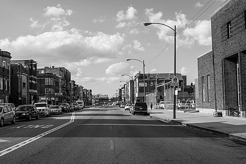 18th Street in black and white, storefronts running both sides under streetlights toward a school crossing sign, parked cars along both kerbs.