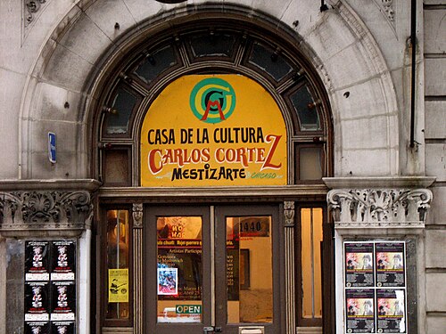 The arched stone doorway of the Casa de la Cultura Carlos Cortez on 18th Street, a hand painted sign over double wood doors and event posters in the windows.