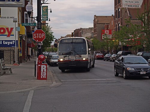 An 18th Street bus stopped at Paulina Street, storefronts with Spanish language signage and a torch style streetlamp under an evening sky.