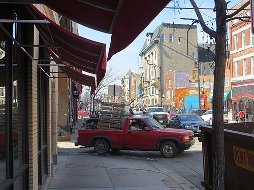 18th Street looking east from under a storefront awning, a mansard roofed corner building and a parked delivery truck down the block.