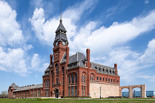 The factory and administration building's brick clock tower and long low wing, arched openings running its length, with a stone archway further along the lawn.