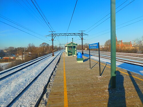 The 111th Street Metra platform on its embankment, snow along the rails and the factory building's clock tower visible past the wires.