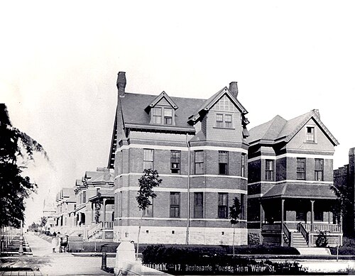 A row of bay fronted brick duplex houses with dormered attics and covered porches, photographed from the sidewalk in 1885.