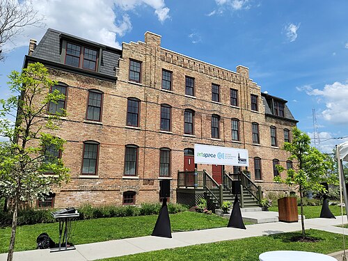 A three storey brick building with arched window heads and a mansard roof, its ground floor entered from a wooden porch and stair.