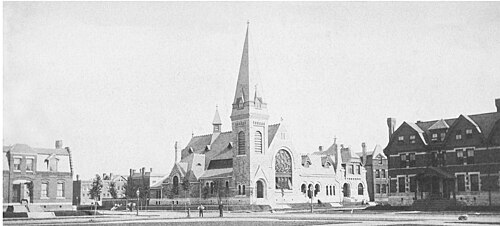 A church with a tall spire and a rose window, photographed from across the green with rowhouses on either side in 1883.