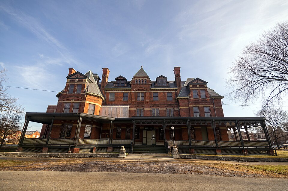 The Hotel Florence's main facade, three storeys of red brick with a steep mansard roof, dormers, and a wraparound porch on iron columns facing an open lawn.