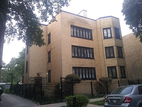 The Kenna Apartments, a three storey yellow brick three flat with ornamented window surrounds and a carved stone entrance, behind an iron fence.