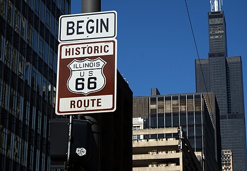 The Route 66 begins here sign on its pole at Adams and Michigan, dark steel towers rising behind it.