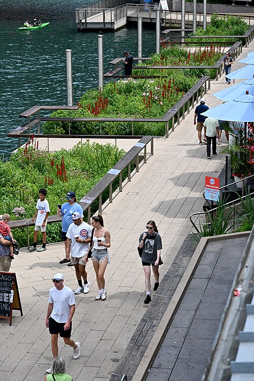 The Riverwalk's paved promenade along the river's south bank, with planters and benches under the office towers.