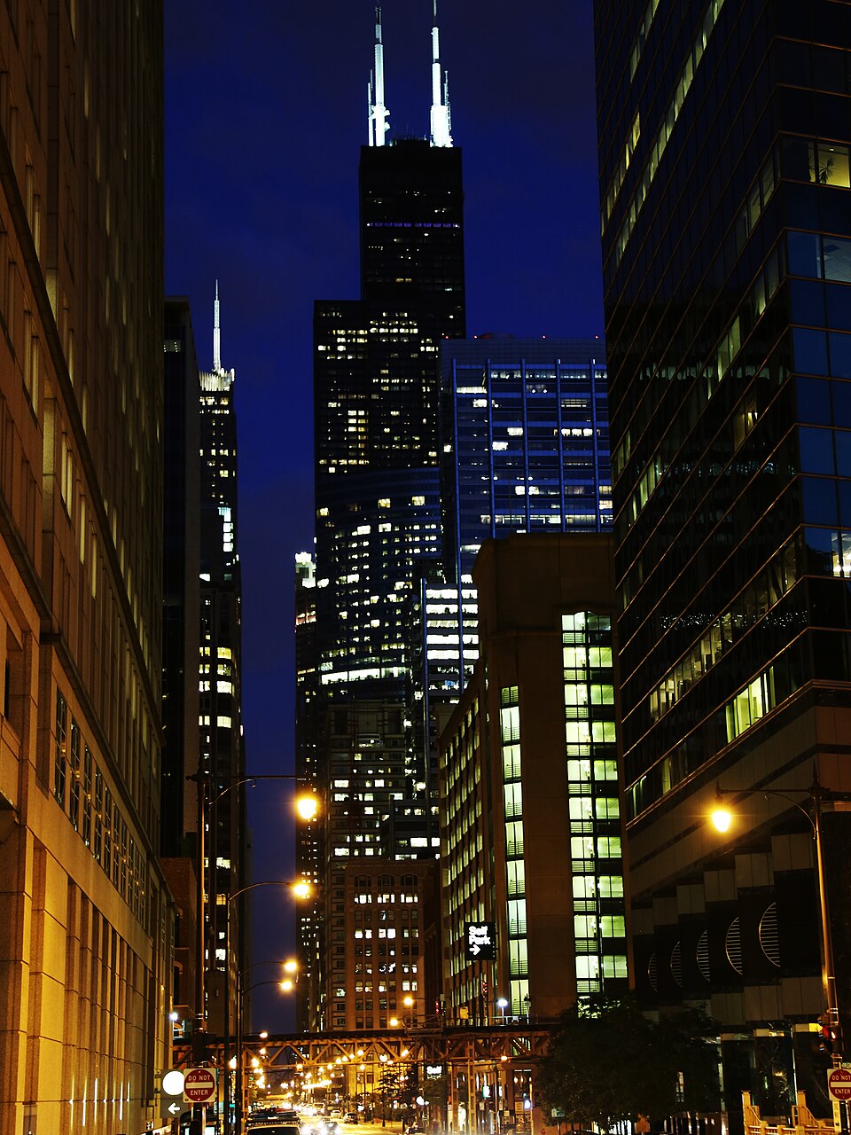 A night view up a canyon of illuminated office towers to Willis Tower, with the elevated train trestle crossing in front.