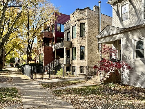 A row of Carmen Avenue houses from different eras, a bay windowed limestone house between a brick two flat with a maroon infill building and a white wood frame porch house.