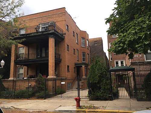 A three storey brown brick apartment building with stacked wrought iron balconies behind a tall iron fence.