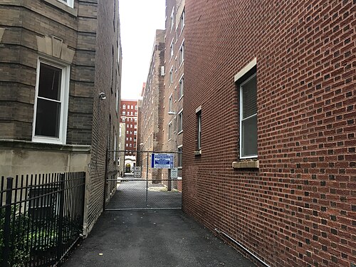 A service alley between brick apartment buildings, chain link gates closing it off at the sidewalk.