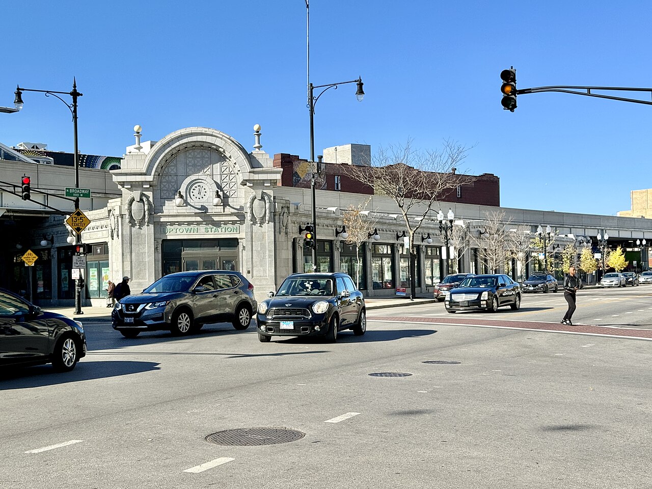 The white terracotta corner building lettered UPTOWN STATION at Broadway and Wilson, its arched clock face over the sidewalk with cars and a crossing pedestrian in afternoon light.