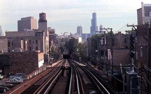 The elevated tracks run south toward downtown, a water tower on a nearby rooftop and the Loop skyline visible past storefronts and utility poles.