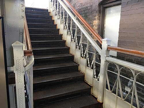 A cast iron station stairway with a wood handrail and pierced Victorian ironwork, brick wall alongside.
