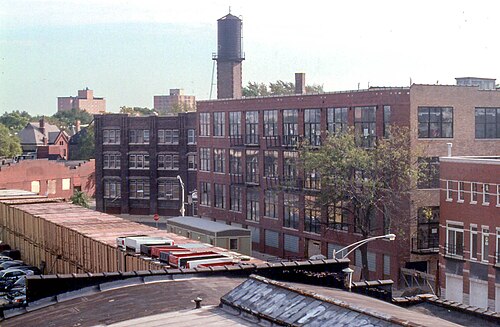 A brick loft building with a water tower on the roof, rail cars on a siding in front, near the edge of the neighborhood.