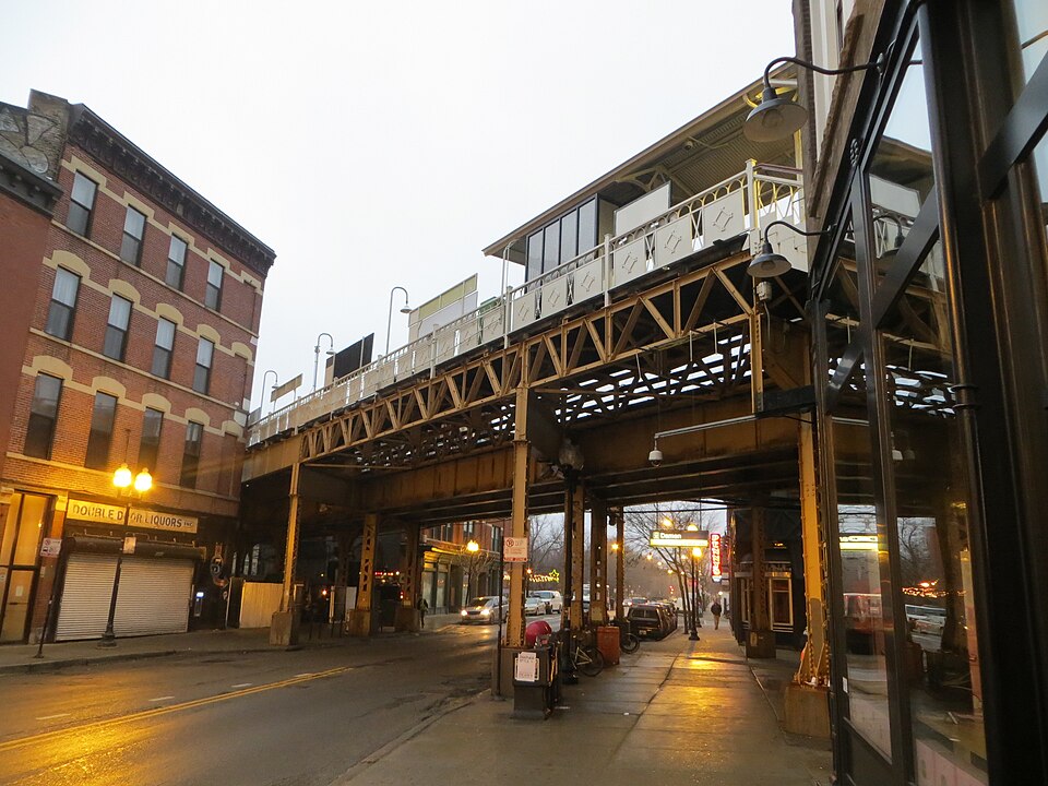The corner storefront a few feet south of six corners, still lettered Double Door Liquors Inc, with the Blue Line's Damen station crossing on steel trusses overhead.