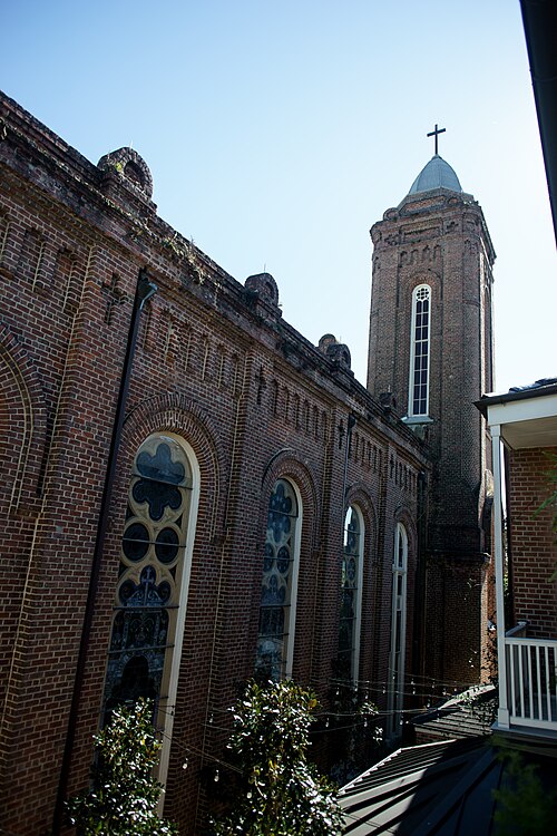 A brick church with a corner bell tower and tall arched windows, seen from a balcony across its courtyard, with string lights hung under the windows.