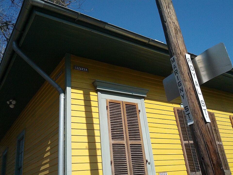 The corner of Royal Street and Marigny Street. A yellow clapboard cottage with brown louvred shutters, the street names on vertical signs, and an older enamel ROYAL plate fixed to the wall.