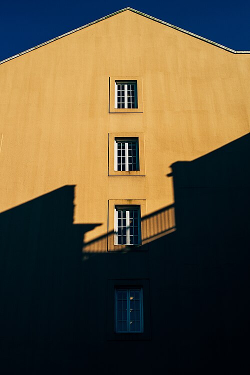 A plain stuccoed end wall with four small windows stacked up it, the shadow of the next building falling across in late sun.