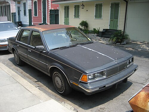 A rusting 1985 Buick Century parked in front of painted Creole cottages with green shutters and a green door.