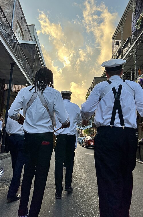 A brass band walking down the middle of a French Quarter street at dusk, seen from behind, galleries over the sidewalk on both sides and the road still wet.