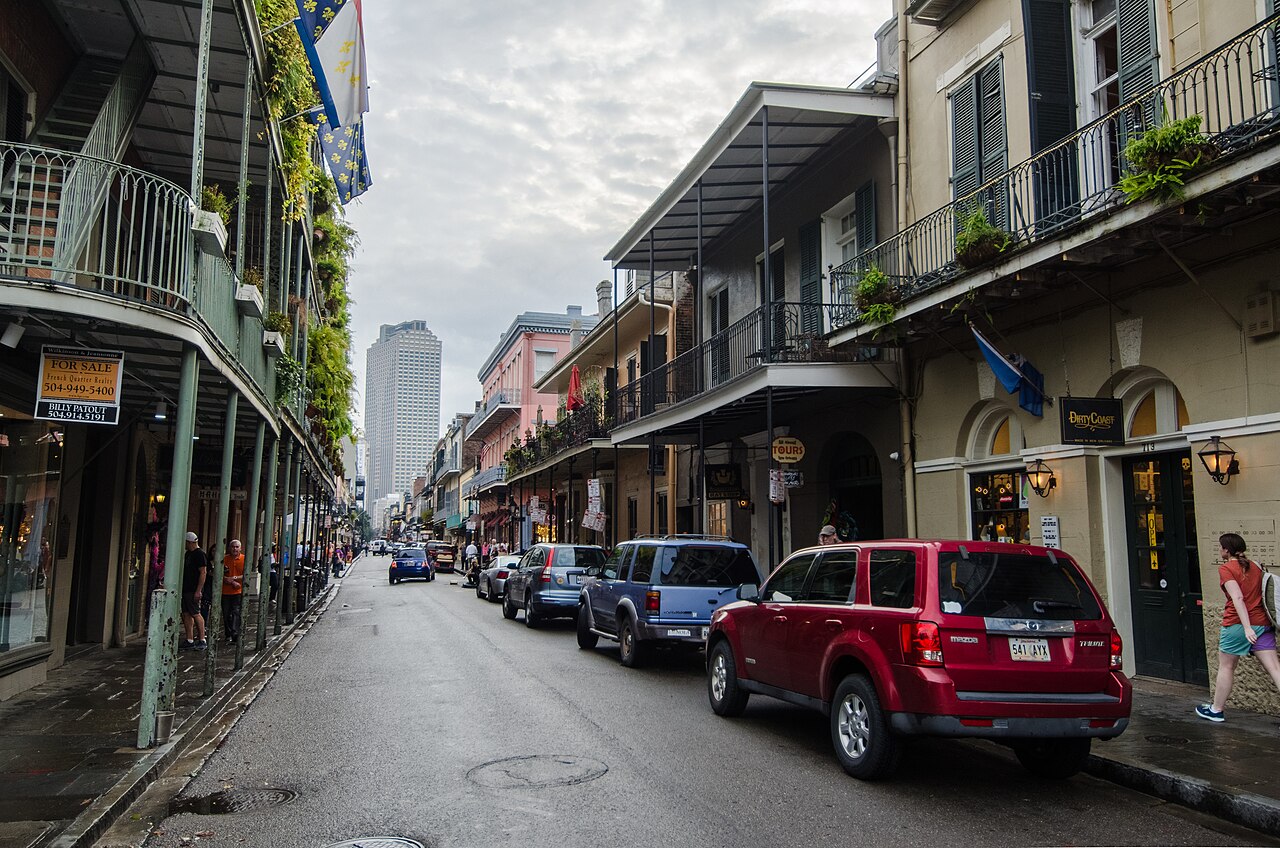 Royal Street between St. Peter and Orleans, looking toward Canal Street. Cast iron galleries over the sidewalk on both sides, cars along the curb, an office tower at the far end.