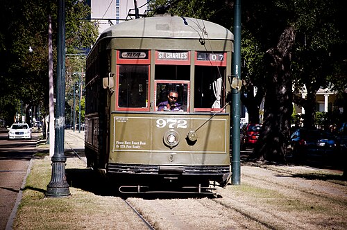 An olive green St. Charles streetcar on the tracks in the grass neutral ground, seen head on with the avenue and its trees on both sides.