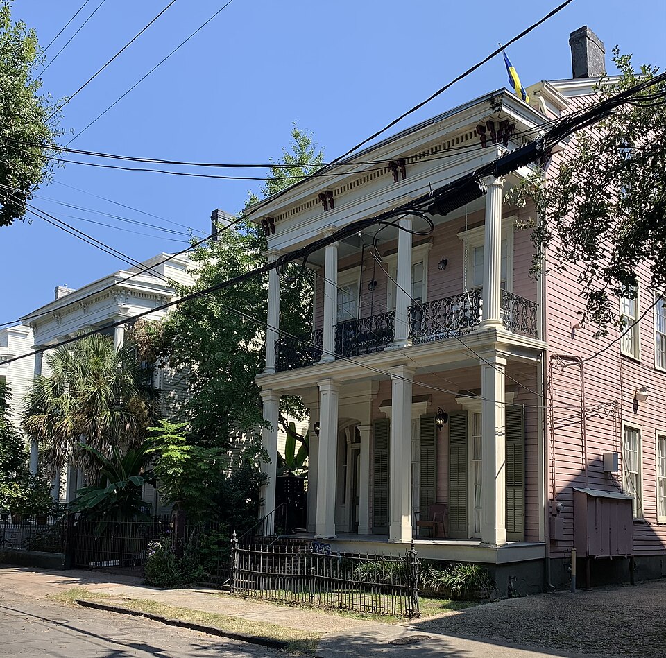 Two double gallery townhouses on the 2700 block of Coliseum Street, pink and cream wood with square columns and cast iron railings, behind a low iron fence.