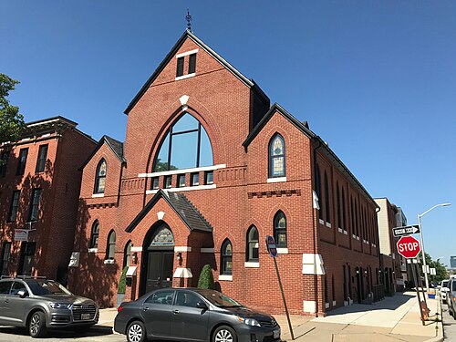 A former Methodist church on South Ellwood Avenue, red brick with a steep central gable, a tall pointed arch window below a louvered vent at the peak, smaller stained glass windows on the side elevation, a rowhouse visible at its side.