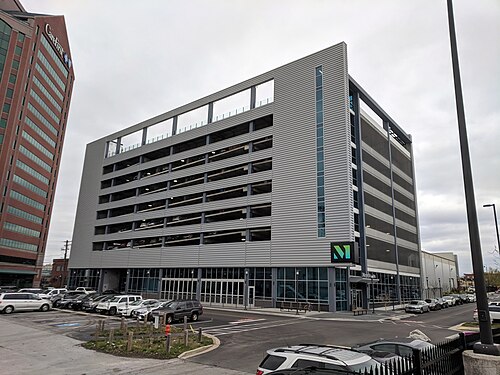 A ribbed grey metal parking garage for the Merritt Clubs gym on Boston Street, cars visible through the open upper levels and the gym's own entrance at street level, the red brick CareFirst tower behind it.
