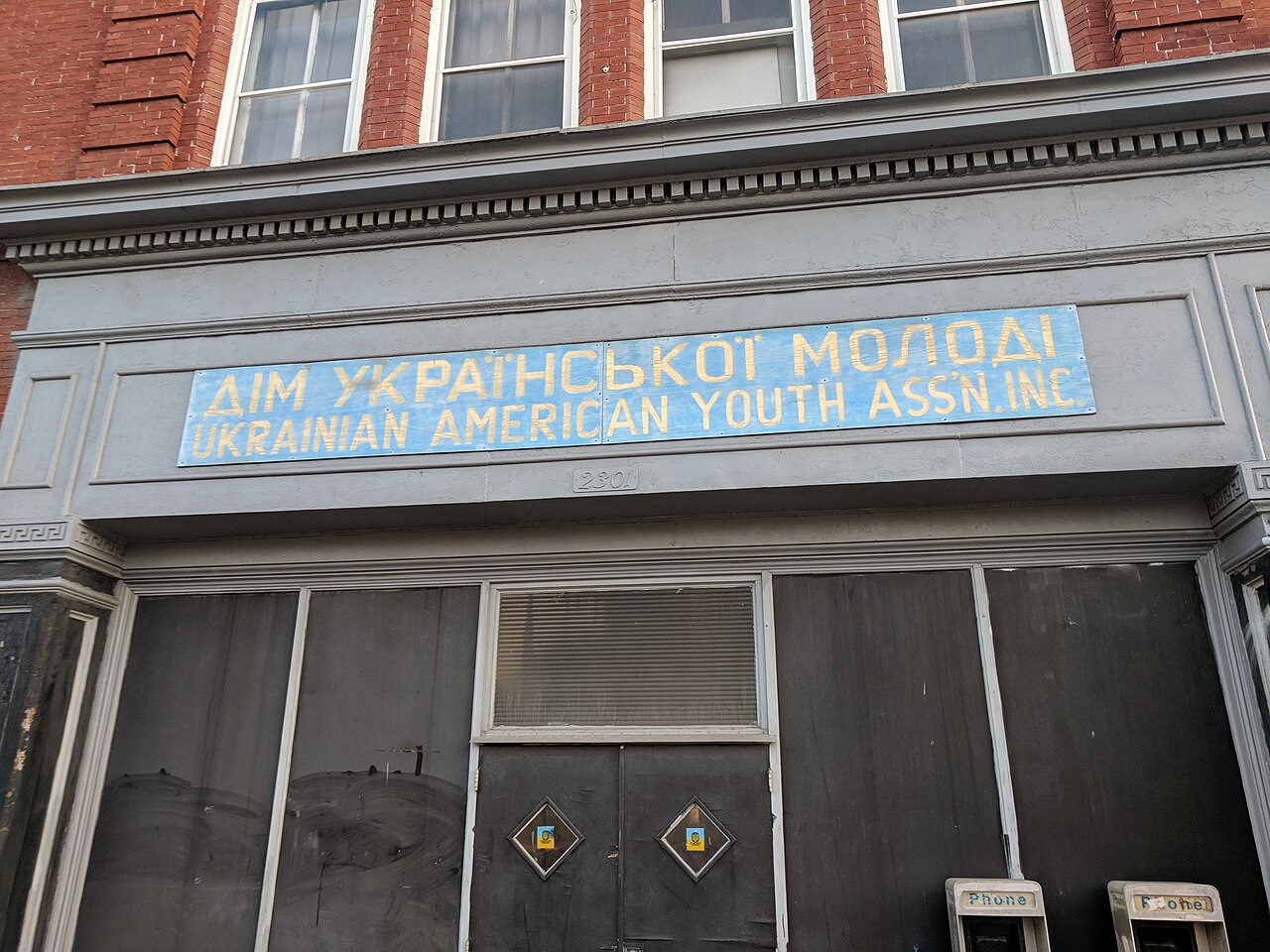 A boarded storefront on Eastern Avenue, a blue sign in Ukrainian and English reading Ukrainian American Youth Assn Inc over its darkened windows.