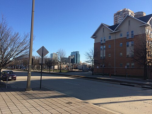 A modern brick apartment building on Key Highway, set back behind a wide paved sidewalk with the downtown skyline visible in the distance.