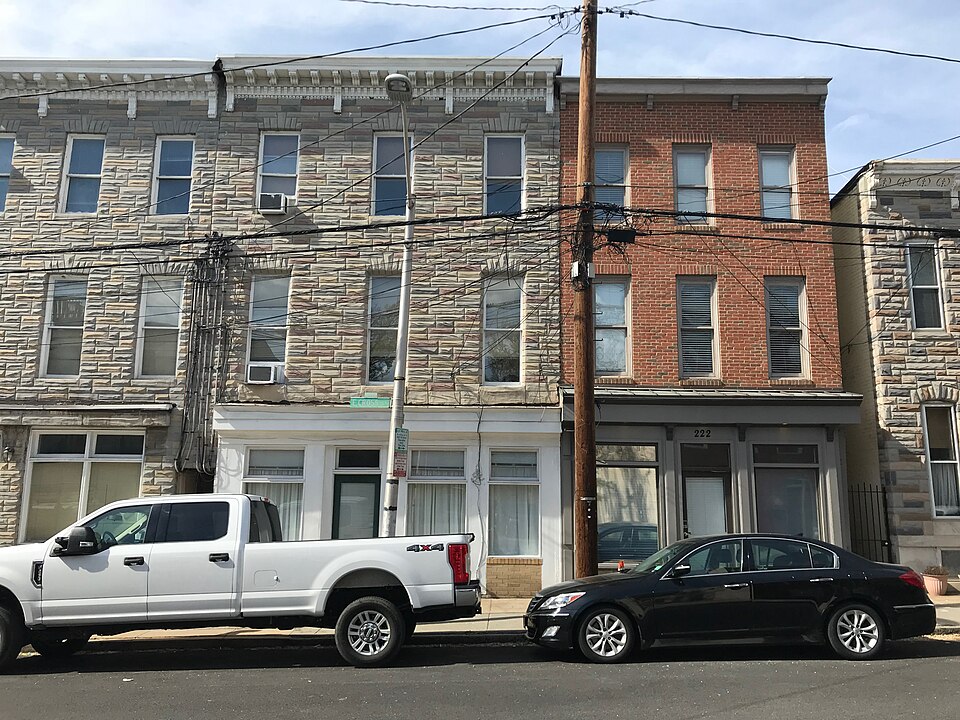 Nineteenth century storefront rowhouses on Cross Street, stone veneer and red brick fronts, three storeys with ground floor shops and cars parked along the kerb. The kind of nineteenth century rowhouse the production used, not the specific one, since the source does not name an address.
