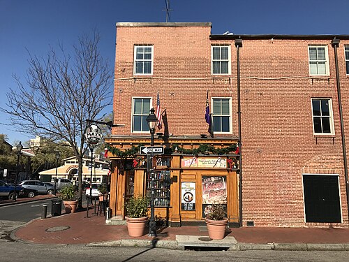 A red brick three storey corner building with a wood pub storefront, potted plants, a hanging sign and a flag beside the door.