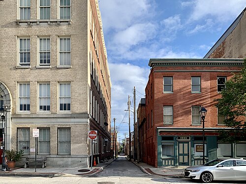 A narrow paved alley between a pale stone building on one side and a red brick rowhouse block on the other, a furniture shopfront at the corner.