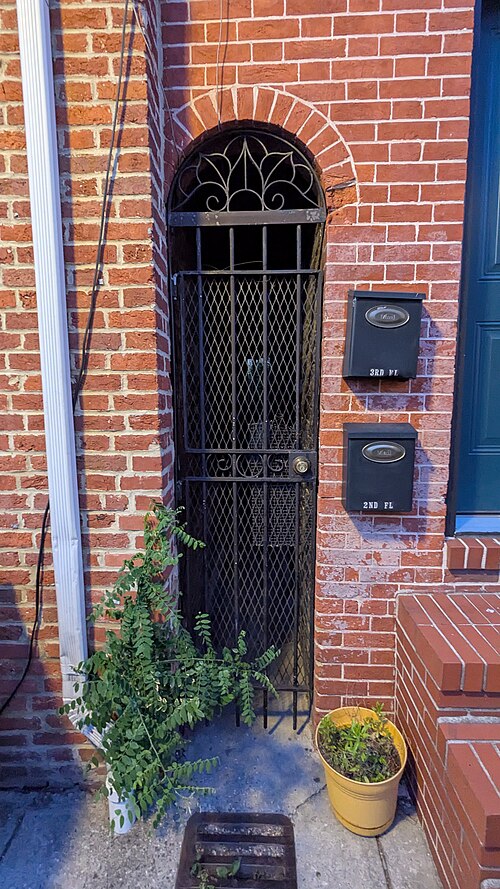 An arched wrought iron security gate set into a red brick rowhouse doorway, floor mounted mailboxes on the brick beside it and a potted plant on the step.