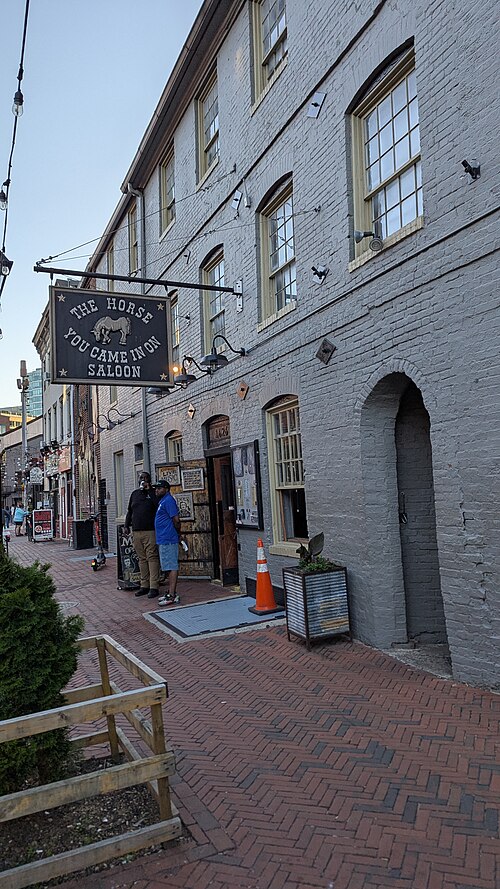 A three storey painted brick tavern building with arched window heads, a hanging sign over the sidewalk and an arched side entrance.