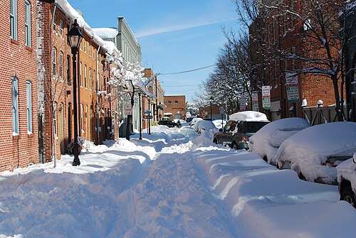A snow covered street between two and three storey red brick rowhouses, cars buried under snow along the curb and a line of cast iron lampposts.