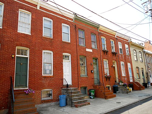 A row of two storey red brick rowhouses with stone steps, iron rails and painted doors, running down a plain sidewalk block.