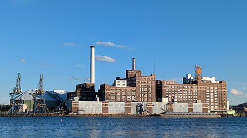 The Domino Sugars refinery across the harbor water, a tall brick industrial block with cranes and a lit rooftop sign, seen from the Fells Point waterfront.