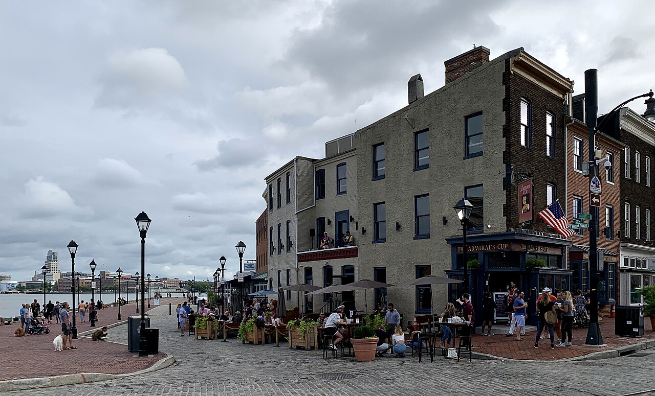 A three storey stucco and brick corner pub with blue trim and a hanging sign, cafe tables under umbrellas along a cobblestone waterfront promenade, brick rowhouses receding down the block.