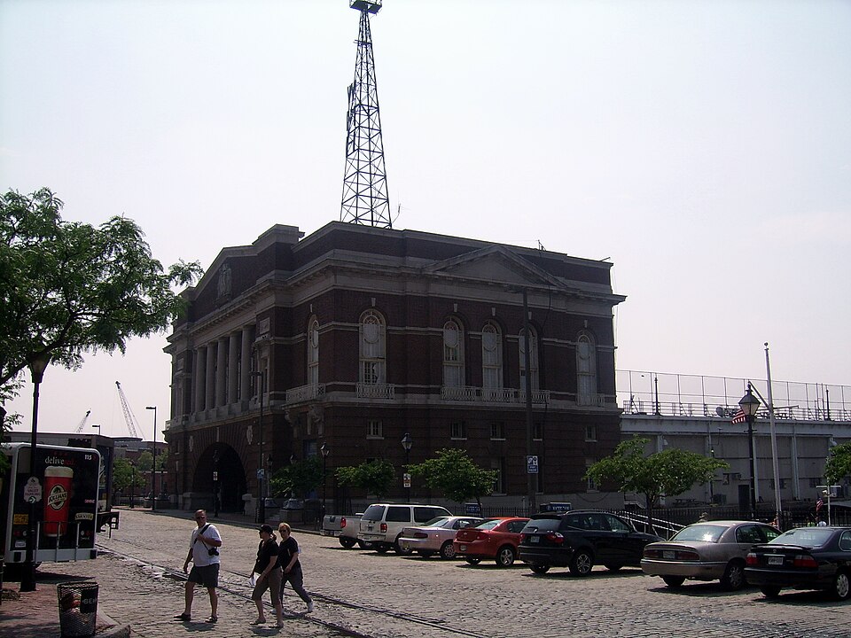 A three storey brick pier building with a columned upper front and pediment, a radio tower over the roof, and cars parked on the cobblestone forecourt below.