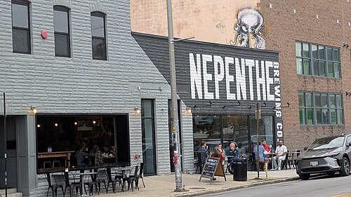 A grey stone textured brewery facade with outdoor seating and painted wall lettering on Falls Road.