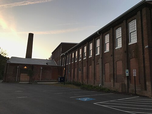 A long red brick mill building with taller rear wings and a tall brick smokestack, on Clipper Mill Road.