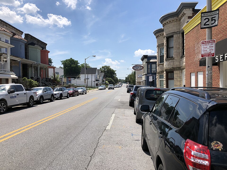 Falls Road looking south from Cox Street, parked cars along a block of stone and brick rowhouses and commercial fronts.
