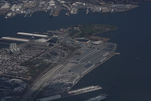 An oblique aerial view of the Locust Point peninsula, showing Fort McHenry's grounds at the tip, the Silo Point tower, container stacks and cranes on the marine terminal, rail yards, and the Fort McHenry Tunnel approach.